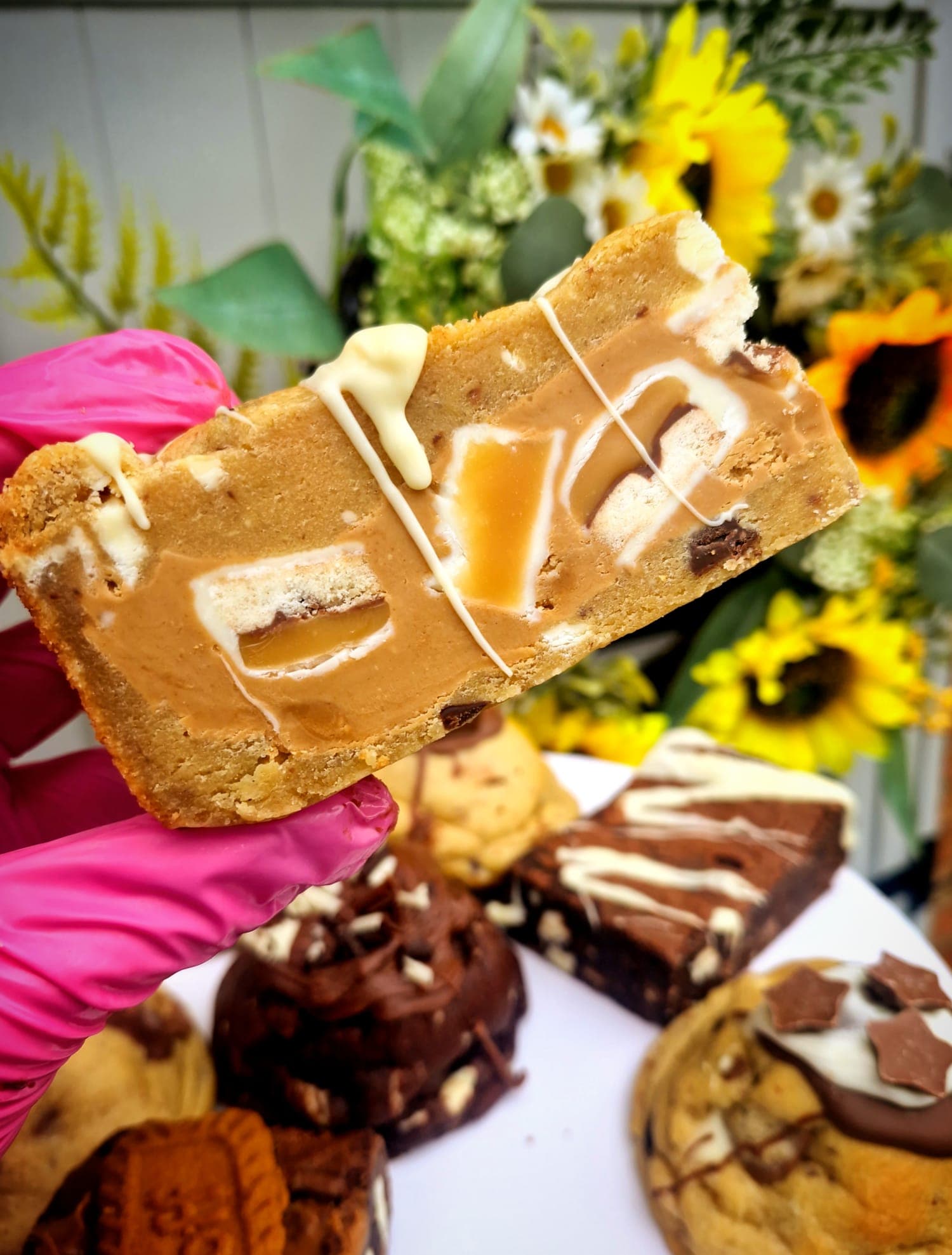 A blondie held up in front of a stand of brownies and stuffed cookies at LoveLea Baking